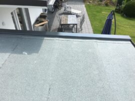 Flachdach mit grauer Beschichtung und einem Blick auf eine Terrasse mit Möbeln im Hintergrund.