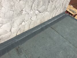 Flachdach mit grauer Abdichtung und strukturiertem Wandbereich im Hintergrund.