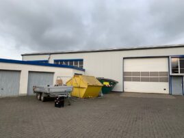 Gelber Abfallcontainer und Anhänger auf einem Parkplatz vor einem Gebäude.