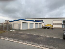 Ein Lagergebäude mit mehreren Garagentoren und einem Anhänger auf einem gepflasterten Parkplatz.