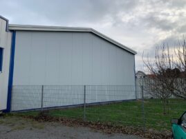 Seitenansicht einer Halle mit grauer Verkleidung und einem Zaun im Vordergrund.
