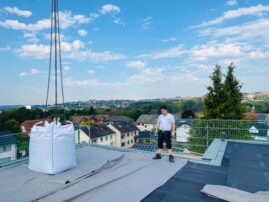 Ein Mann steht auf einem Flachdach neben einem großen Sack mit Baustellenmaterial und blickt in die Ferne.
